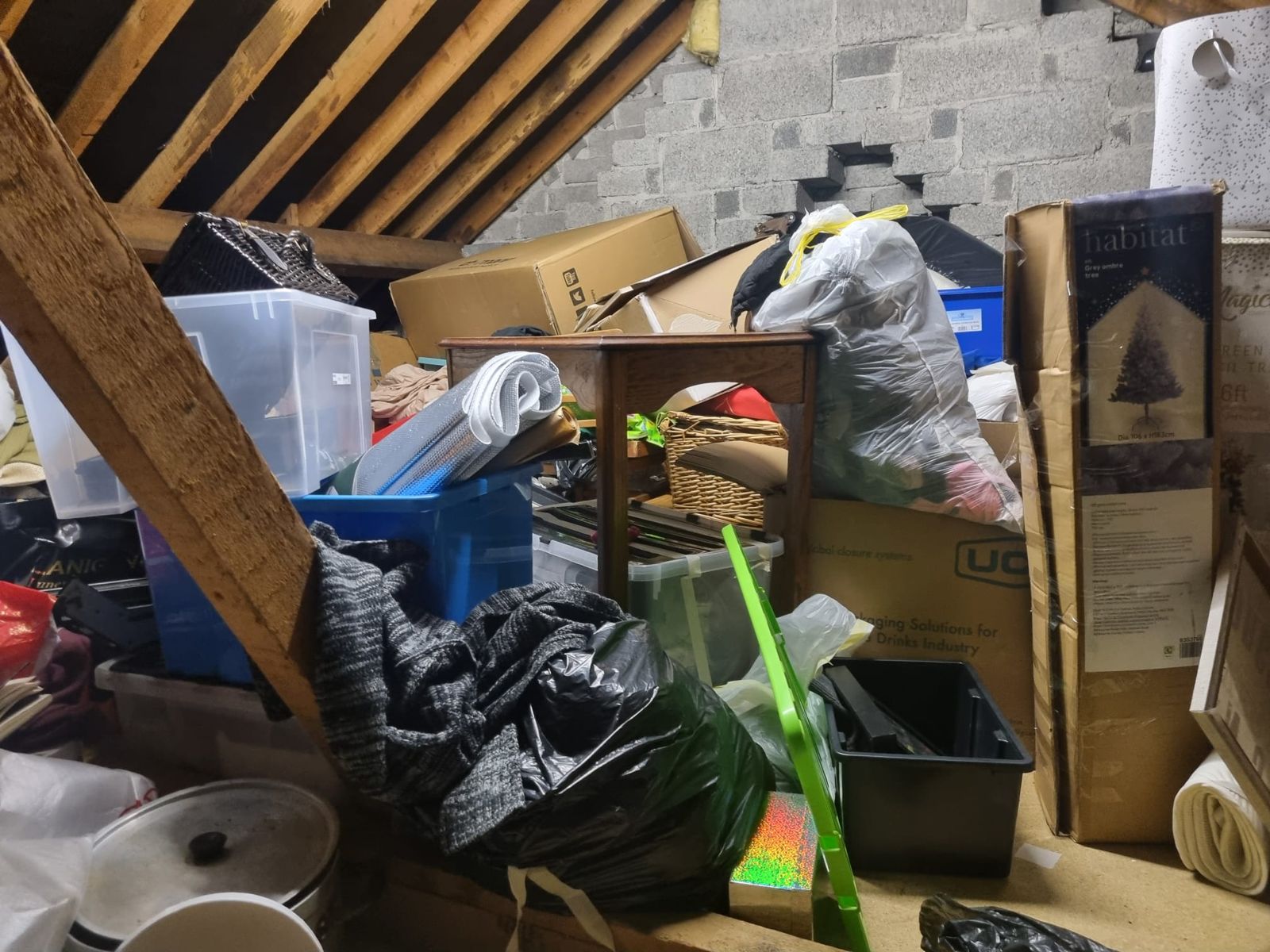 An attic under the roof beams, full of boxes, bags and a boxed Christmas tree
